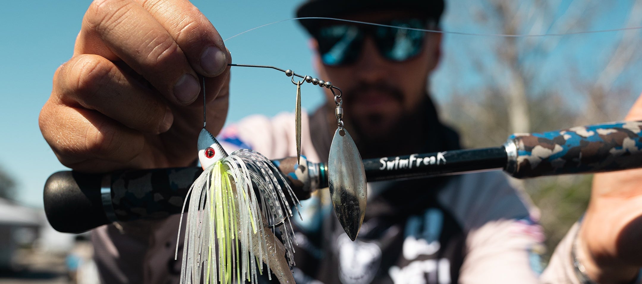 Person holding a fishing rod with a Bassman lure, wearing sunglasses and a cap, outdoors.